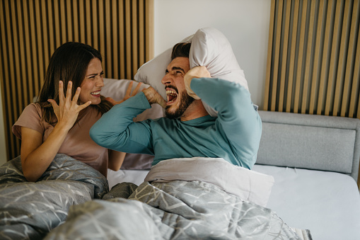 Annoyed adult woman and man in bed suffering neighbor noise at home. The couple has an unhappy facial expression closing their ears with pillows while lying in bed.