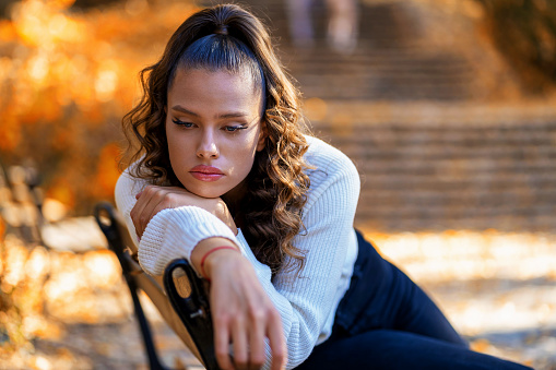 A girl with long hair is sitting on a bench looking pensive. She is in a park and is dressed casually. She is resting her head on her hand.