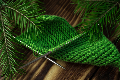 Started knitting on knitting needles, with yarn on a rustic wooden table, with green branches of a Christmas tree. We knit warm clothes with our own hands for cold winters and frosts during the crisis