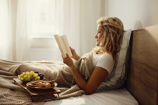 Young smiling woman reading a book during morning in a bed.