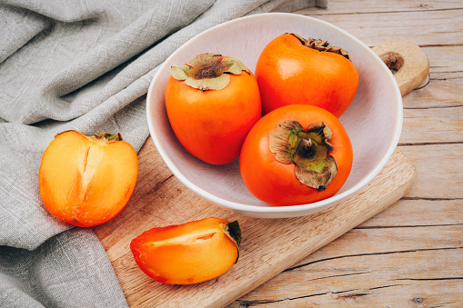 Fresh ripe persimmons in white bowl on cutting board and napkin on wooden table. Healthy vegan food concept. Top view.
