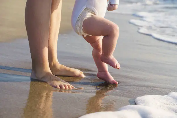 Photo of Baby walks on the sea beach and learns to walk with his mother. First steps, parental care and love concept