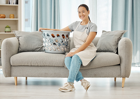 Cleaning laundry, service and home woman working to clean basket of clothes, living room sofa and house linen in an apartment. Portrait of Asian cleaner staff in lounge doing housekeeping with smile