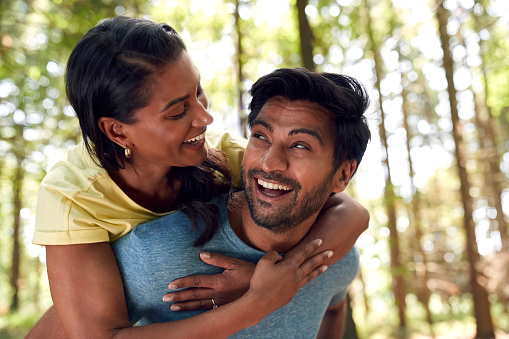 Portrait Of Couple Hiking Or Walking Through Woodland With Man Giving Woman Piggyback