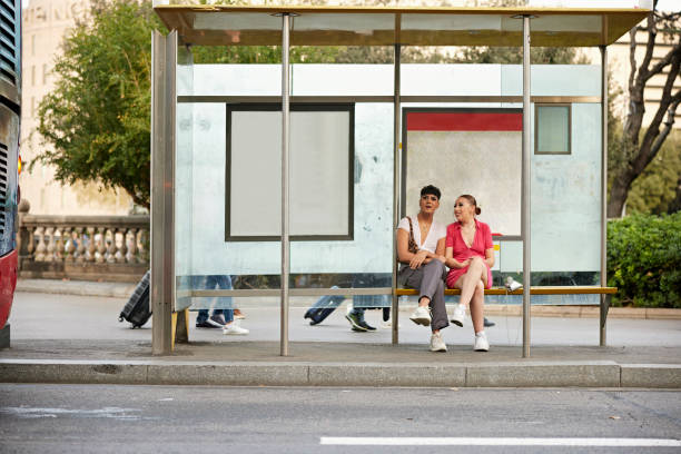 Young people waiting for public transportation in Barcelona Full length front view of early 20s woman in pink dress talking with gay 18 year old friend as they sit and wait inside bus shelter. bus shelter stock pictures, royalty-free photos & images