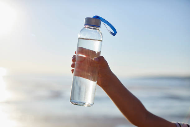 Clean drinking water in glass bottle held by boy Hand of boy holding glass bottle against sky. Male child is at beach during sunset. blue reusable water bottle stock pictures, royalty-free photos & images