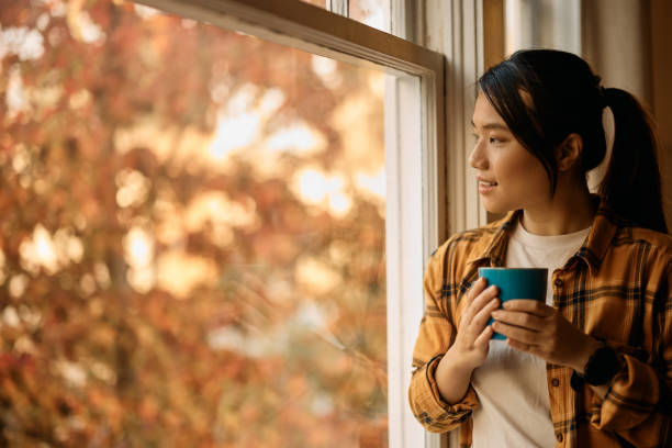 istock Young Asian woman having cup of tea while standing by the window and looking through it. 1439947458