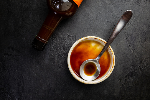 Worcestershire sauce in a bowl with spoon and bottle over black background, top view