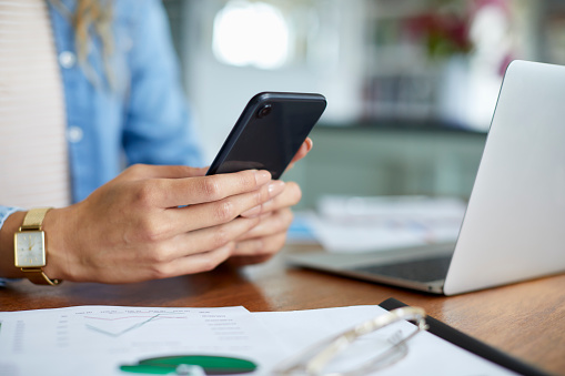 Hands of businesswoman holding mobile phone by laptop at desk. Female entrepreneur is surfing net on phone. She is text messaging.