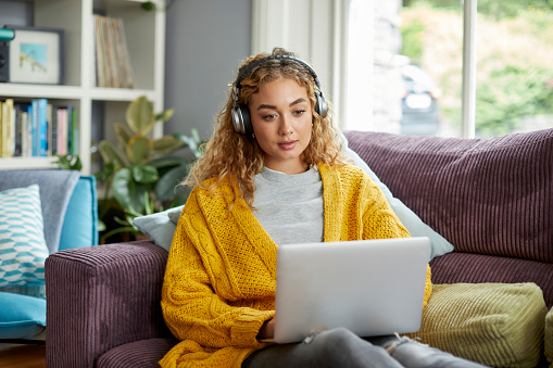 Beautiful blond woman sitting on sofa using laptop at home. Young female is listening to music through wireless headphones. She is surfing on internet.