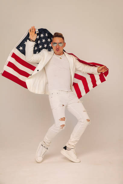 istock Confident successful handsome young man in elegant white classic suit and sunglasses dancing with us flag at grey background, looking at camera. Studio shot of show presenter, dancer, model concept 1439949635