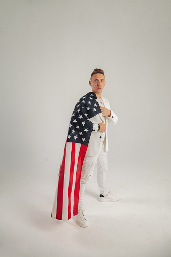 Confident successful handsome young man in elegant white classic suit standing with us flag at grey background, looking at camera. Studio shot of show presenter, dancer, model concept. Copy text space