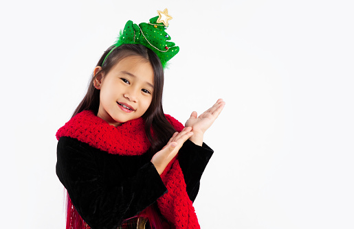 Asian kid girl long hair style in christmas theme costume and christmas tree headband pointing hand and smiling looking at camera white background.