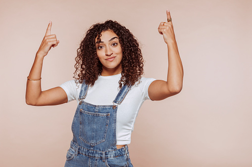 Portrait of young arabic modern woman with curly hair, wears denim overalls, points with two hands and fingers up. Studio portrait isolated over beige background.