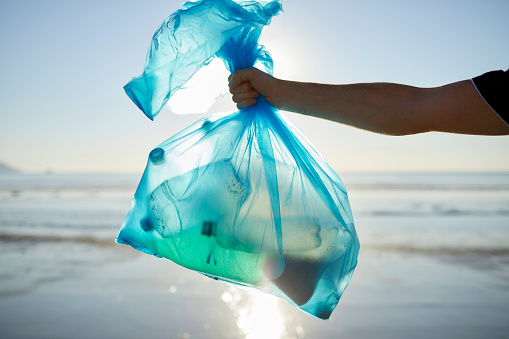 Hand of male environmentalist holding garbage in plastic bag. Man is collecting abandoned waste against sea. He is cleaning up beach.