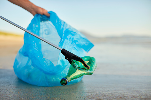 Male volunteer's hand holding blue plastic bag collecting waste. Environmentalist is collecting abandoned water bottle with litter picker. Activist is cleaning beach.