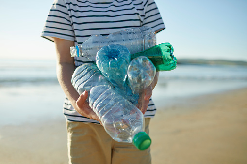 Midsection of boy holding empty plastic water bottles at beach. Male environmentalist is standing on shore. He is doing beach clean up.