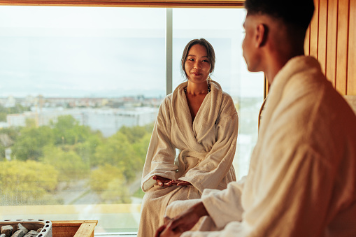 A young mixed race couple is in a sauna in their hotel. They are sitting in their bathrobes as the sauna's window has a view of the city.