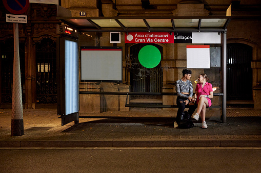 Full length front view of early 20s woman in pink dress sitting under public transportation shelter with gay teenage friend and talking.