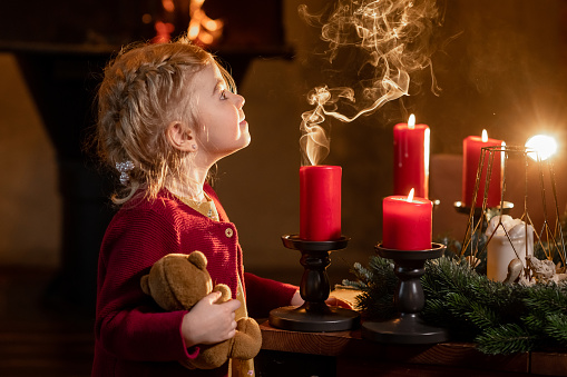 Cute child girl blow candles on Christmas and New Year eve evenings at cosy fireplace background home. Concept of family and miracle.