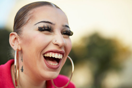 Close-up of 20 year old with hair slicked back wearing pink dress, large hoop earrings, false eyelashes, looking away from camera and laughing.