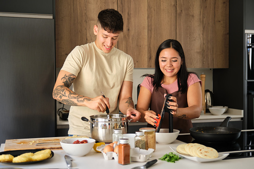 Two young hispanic people smiling while mashing potatoes and grating tomato at kitchen.