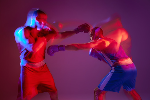 Unfocused effect portrait of two professional boxers boxing isolated on purple background in neon light. Concept of sport, competition, training, energy. Copy space for ad, text