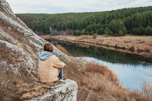 Young woman in beige jacket sitting on rock and looking at the view of the river and forest in autumn seasonal neutral colors of nature tourism copy space
