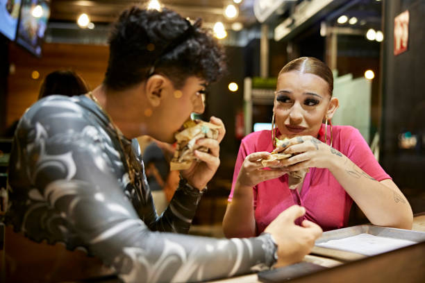 Candid portrait of hungry young people eating and talking Waist-up view of early 20s woman in pink dress looking at gay teenage friend as they sit in Barcelona fast food restaurant after enjoying night out together. crossdressing clothes stock pictures, royalty-free photos & images