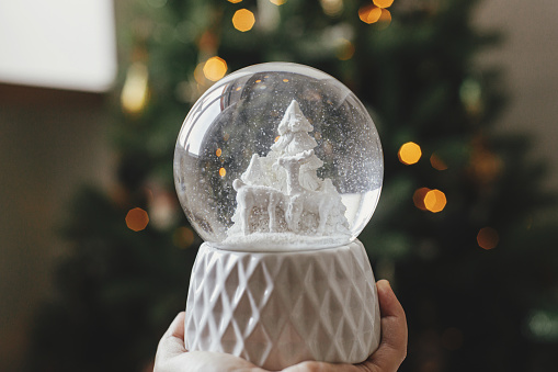 Stylish christmas snow globe in hand on background of christmas tree in lights in festive decorated boho room. Person holding snowy snow globe. Merry Christmas! Atmospheric winter time