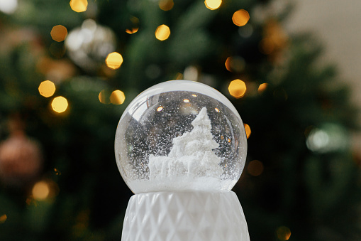 Merry Christmas! Atmospheric winter time. Stylish christmas snow globe on background of christmas tree in lights in festive decorated boho room. Snowy white snow globe against lights bokeh