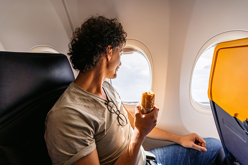 Handsome young man looking out the window of an Airplane and eating a sandwich.
