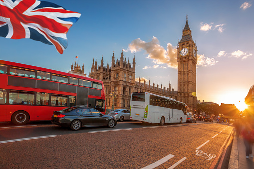 Big Ben with buses on the bridge and flag of England against colorful sunset in London, England, UK