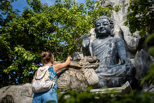 Young tourist placing incense on a Buddha image. Tourist showing respect towards the Buddhist religion. Young woman placing incense at the feet of huge buddha statue