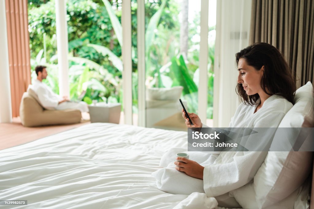 Woman enjoying the lazy morning on her luxury vacation Young woman is using a phone while drinking coffee in bed on her vacation in a luxury villa. 25-29 Years Stock Photo