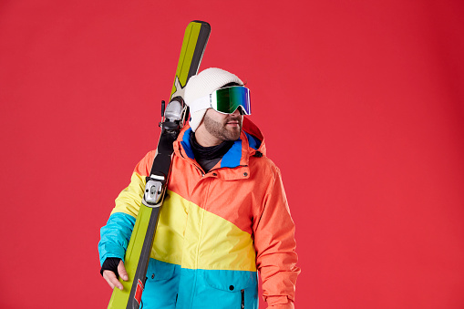 Boy wearing a hat, winter clothing and snowboard goggles looking to the side while holding skis on a red background.