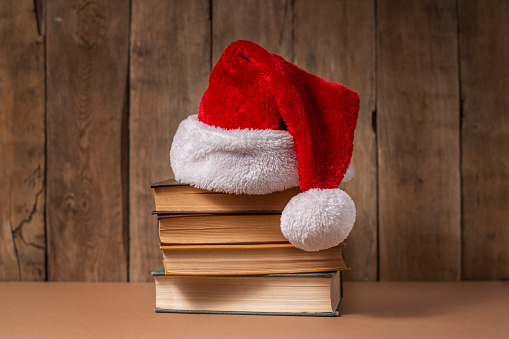 Stack of books and Santa Claus hat on wooden background.