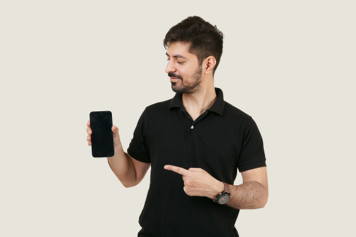 Happy Asian man looking and pointing towards the mobile phone. man holding a mobile phone with a black screen. isolated man holding a cell phone with plain beige background. App advertisement.