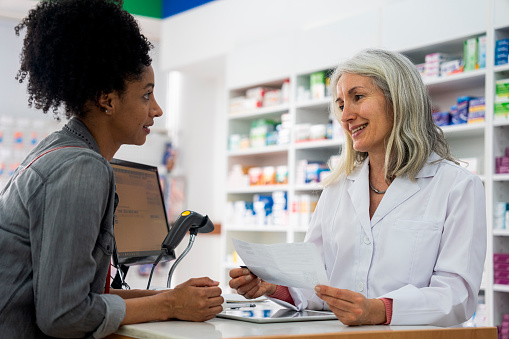 Mid-shot of mature female pharmacist taking an African-American woman's prescription order while standing behind pharmacy's counter