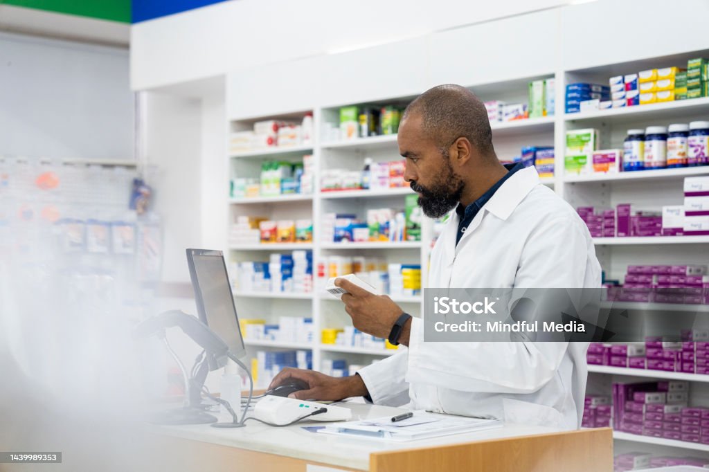 A male African-American pharmacist takes notes on medication inside pharmacy premises Mid-shot side view of male African-American pharmacist writing report on computer inside pharmacy 35-39 Years Stock Photo