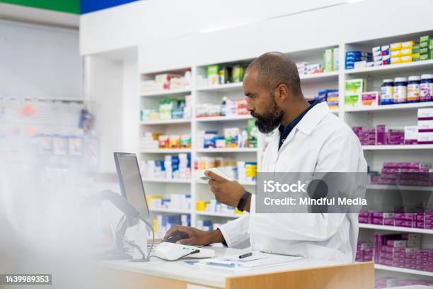 A Male Africanamerican Pharmacist Takes Notes On Medication Inside Pharmacy Premises Stock Photo - Download Image Now
