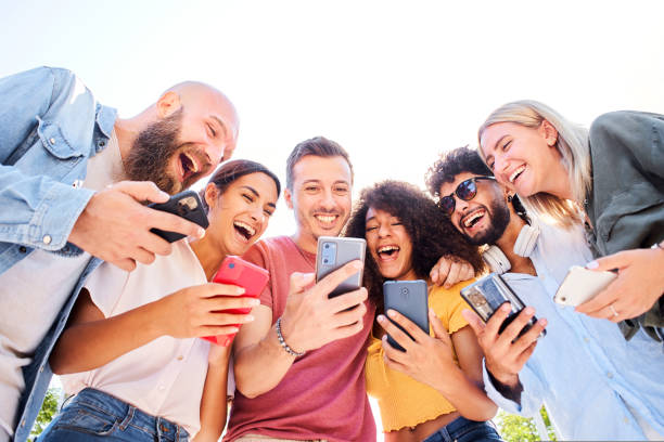 Low angle of Multiracial happy group of friends using phone and smiling together outside. People addicted to technology and social media stock photo
