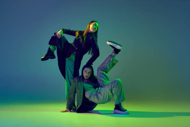 istock Stylish young girls in casual clothes dancing experimental style dance over gradient blue-green background at dance hall in neon light. Youth culture 1439990922