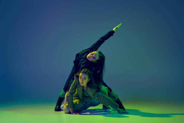 istock Weird. Stylish young girls in casual clothes dancing experimental style dance over gradient blue-green background at dance hall in neon light. Youth culture 1439990946