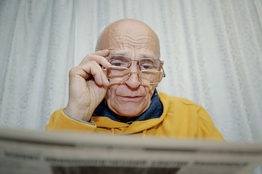 Eccentric grandpa reading newspaper and holding glasses pair with hand. Old people lifestyle.