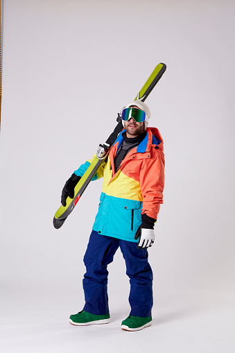 man in snowboard goggles and snow gear holding skis in one hand while looking at the camera on a white background.