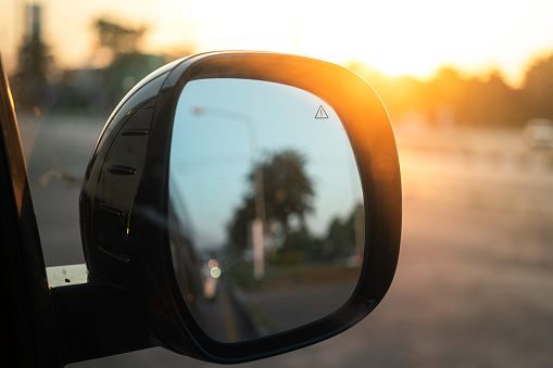 Close-up at side mirror of the car with orange sunlight and flare effect as blurred background. Road trip travel concept. Photo contained noise and high contrast ration due to backlit scene.