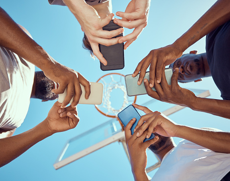 Men in a circle with phones networking on social media while standing on a basketball court. Sports group doing research on team strategy, collaboration and skill together with technology from below.