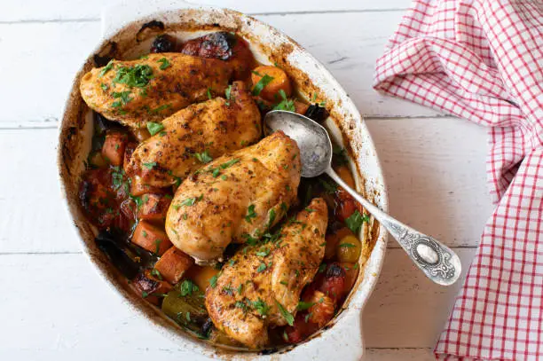 Oven roasted or baked chicken breast with mixed vegetables. Cooked in a casserole dish and served hot on white wooden background from above. Healthy eating meal for fitness, dieting or low carb nutrition