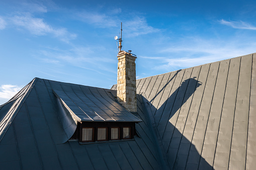 Hipped roof with roof window (dormer) and covered with grey metal seam sheet. Stone chimney with cap and little transmitter. Blue, cloudy sky in the background.